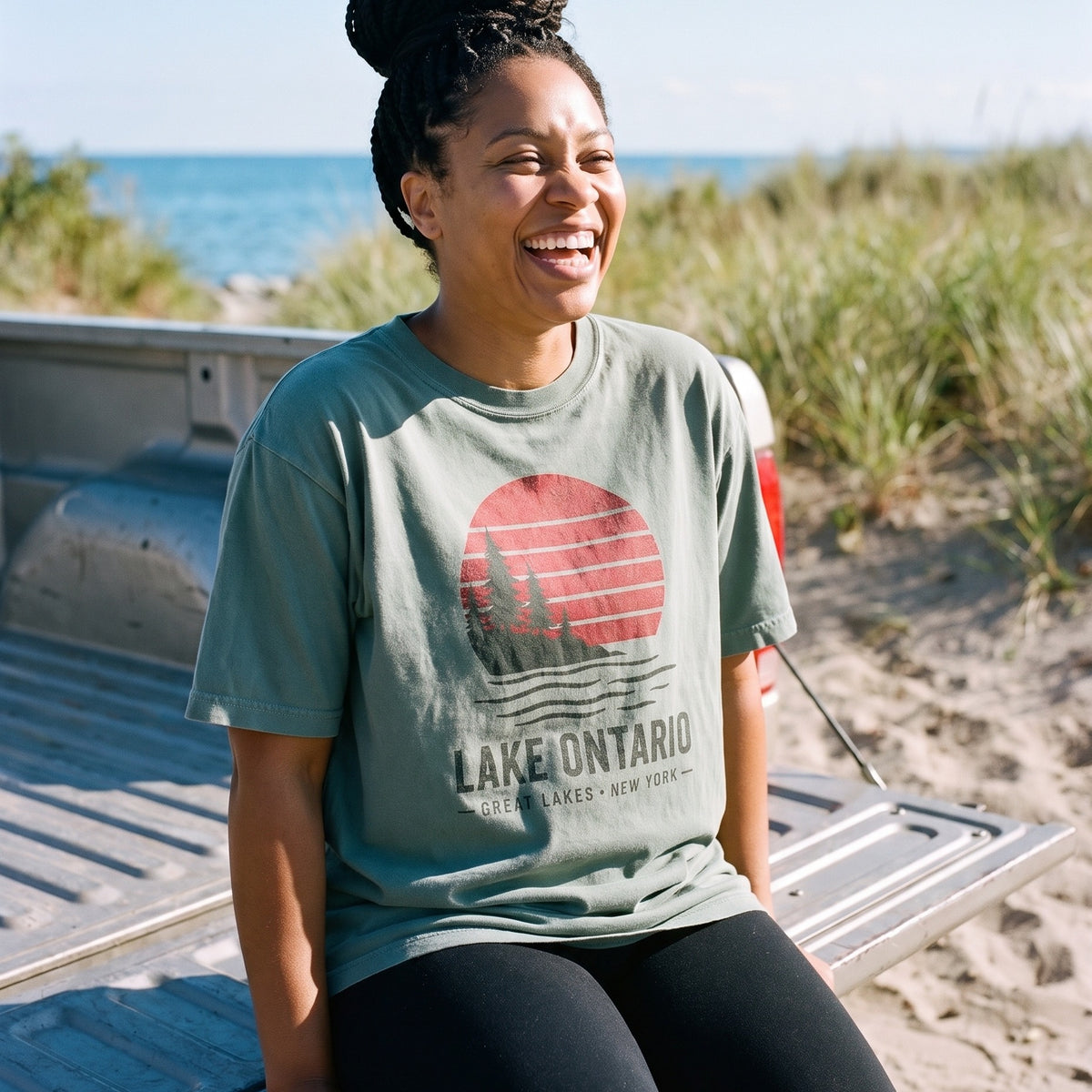 Lake Ontario Sunset tee in Faded Eucalyptus, sitting on a truck tailgate at the beach